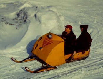 This is an image of an early model Ski-Doo with two riders on it.  The Ski-Doo is seen driving along a snow-covered path.