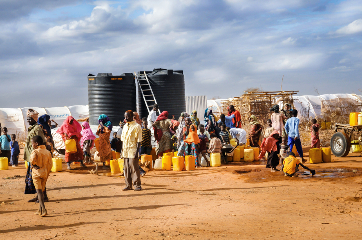 This is a photograph of people lining up for water.  Many are carrying jugs and containers and it appears to be a desert like setting.
