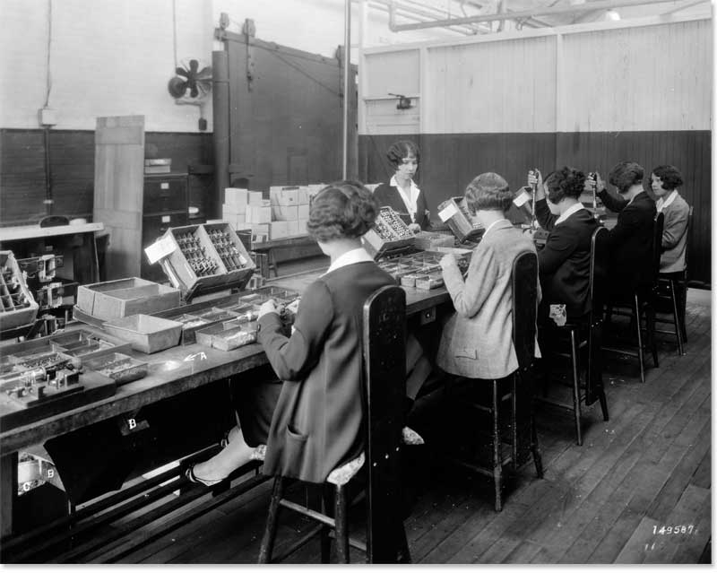 There is a row of five women in an old-fashioned factory room. They are participating in a relay test.