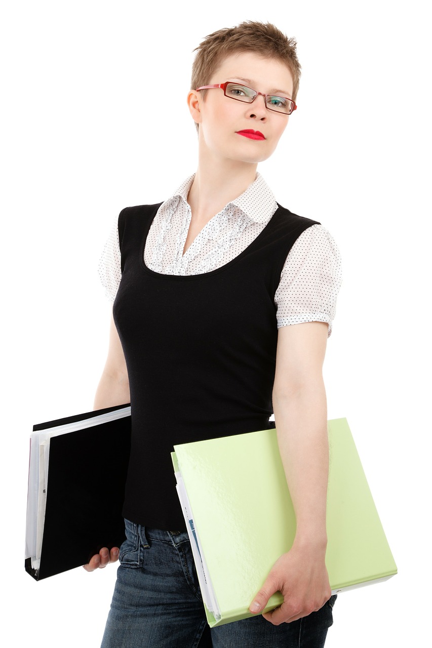 This is an image of young woman wearing jeans, a white T-shirt and black vest carrying a black binder in one hand and file folders in the other.