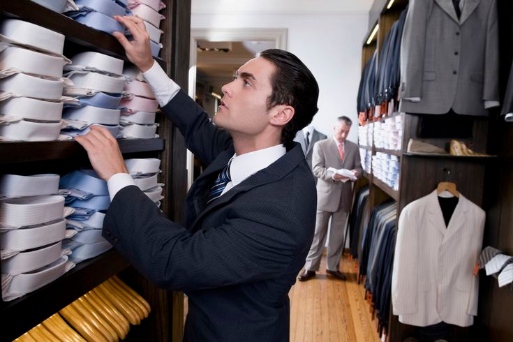 This is an image of a male clothing store worker organizing the dress shirts on a shelf.