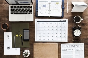 This is a photograph of a desk with a laptop, cellphone, dayplanner, calendar, clock, pen, binder, plant, file folder and new