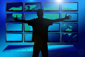 This is a graphic of the outline of a person embracing a wall of computer screens showing a map of the world. There is a keyb