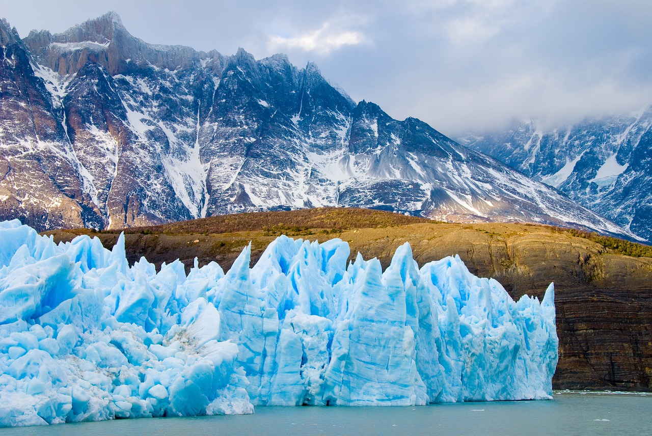 This is a photograph of a glacier in the water in the foreground, and in the background is a row of mountain peaks dusted with snow. 
