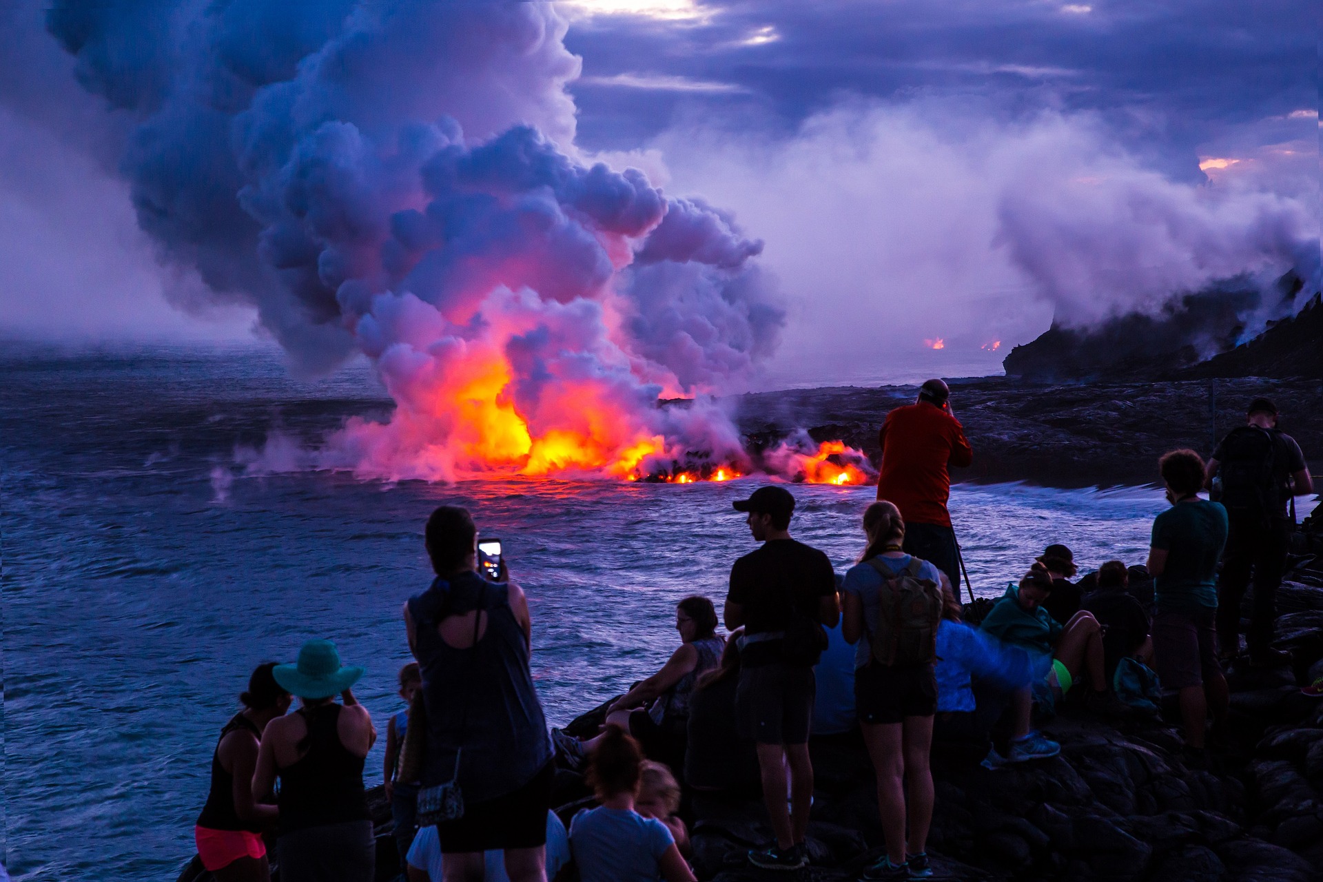 This is a photograph of a group of tourists watching and photographing lava pouring off a coastline into the sea at dusk. 