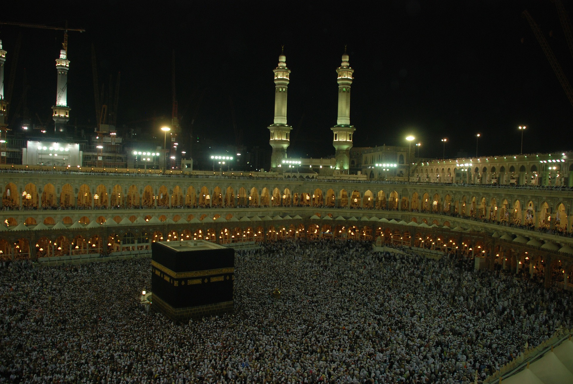 This is a photograph taken at night of thousands of people inside a massive mosque surrounding a black stone building. In the background, the minarets of the mosque are visible.