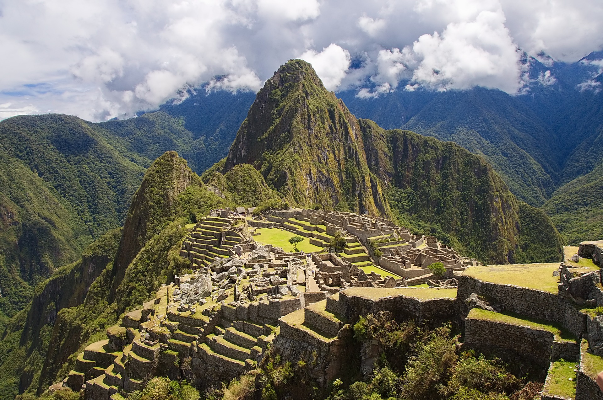 This is a photograph of the ruins of a city on a mountain peak. All of the surrounding mountains are lush with vegetation.