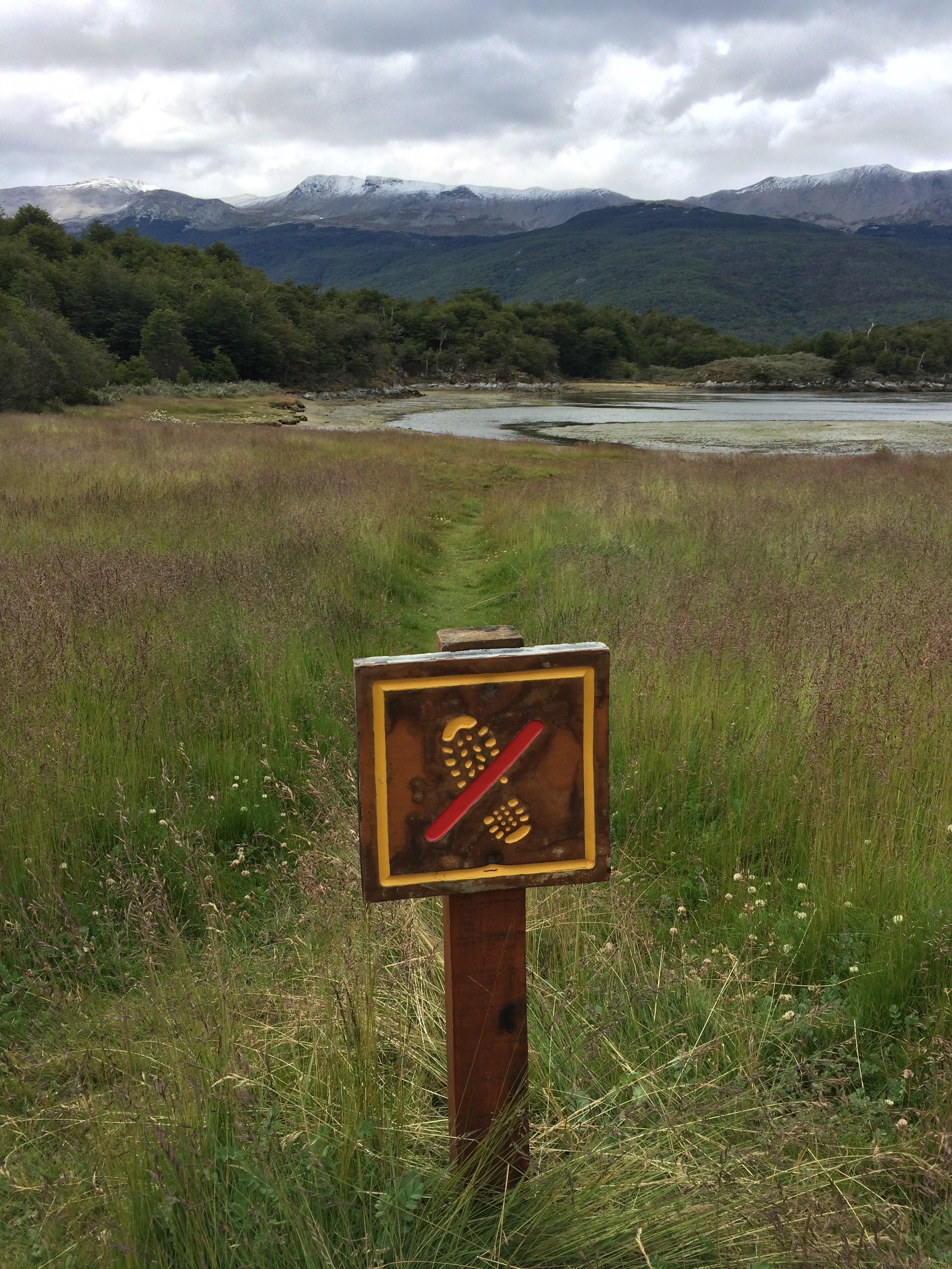 This is a photograph of a meadow with a clear walking path through it which is blocked by a sign showing a hiking boot print and a red slash across it.