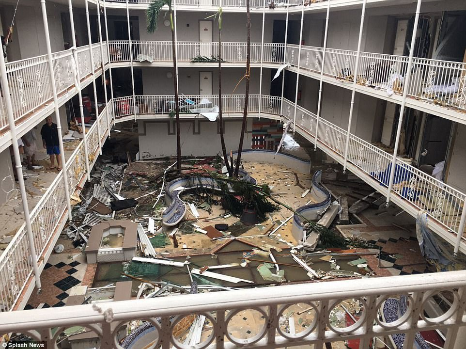 This is a photograph of the inner courtyard of a hotel. There is debris on the balconies and the ground, and the tops ripped off the palm trees.