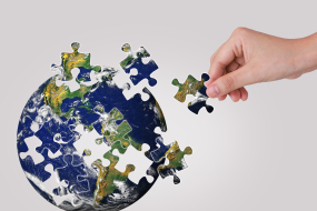 A globe is broken into puzzle pieces. A hand is reaching towards the globe to insert a puzzle piece.