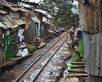 Shacks are extremely close to a train track. A couple of people are walking along the track. The housing is made of found, or scavenged materials. These shelters do not appear to be weathertight.