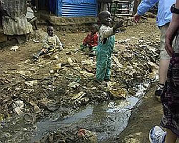 Three small children are in the dirt alleyway. Two are sitting in the dirt and the third is standing on accumulated trash at the edge of some running water. 