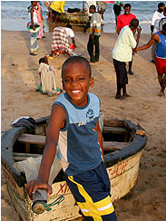 This is a photograph of Uivas Michael.  He is sitting on a rowboat.  There are several people in the background carrying on with their day. Beyond the people is a body of water.