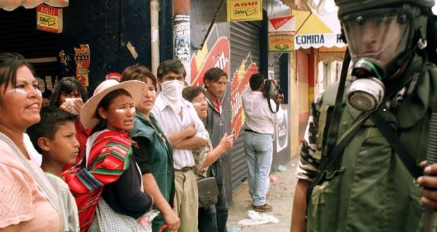 This photograph shows people standing in front of shop in a narrow street.  A police officer in a gas mask stands in foreground.