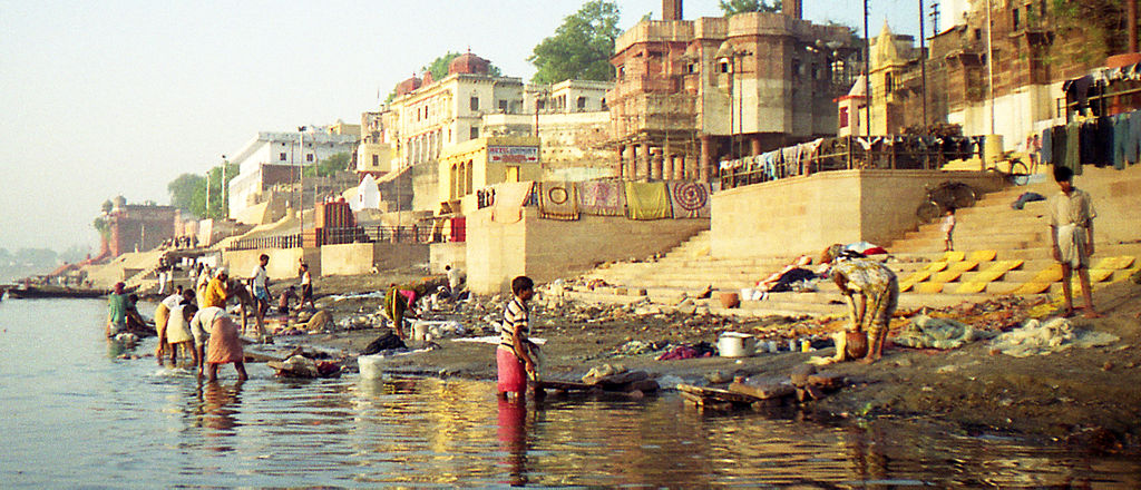 This is a photograph of the Ganges river flowing through the city of Varanasi.  In the background are houses and buildings constructed to the edge of the river. In the foreground, people are washing in the water and drying clothes on the bank.