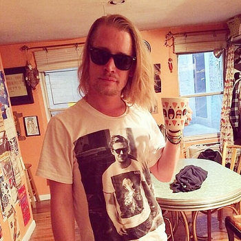 This is a photograph of actor Macaulay Culkin. He is standing in a kitchen, wearing sunglasses and has long, blond hair. He is wearing a t-shirt with an image of actor Ryan Gosling, wearing a t-shirt with an image of Culkin.