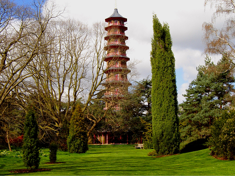 A photograph of a well-manicured lawn and trimmed hedges. In the background, a tall, thin pagoda of about 10 storeys rises. It is painted red with blue tiles. It does not look exactly like a traditional pagoda.