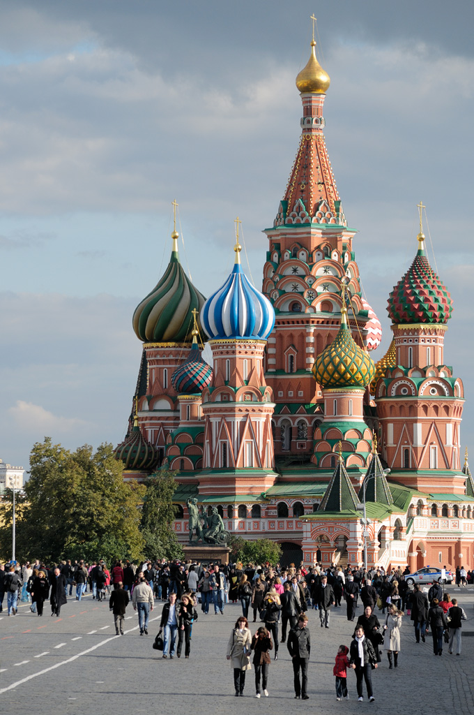 This is a photograph of a church in Moscow. There are 7 onion-shaped domes in various sizes and colours on the top of highly decorated pillars in a pink stone. All are decorated with a variety of stripes, dots, and arches.