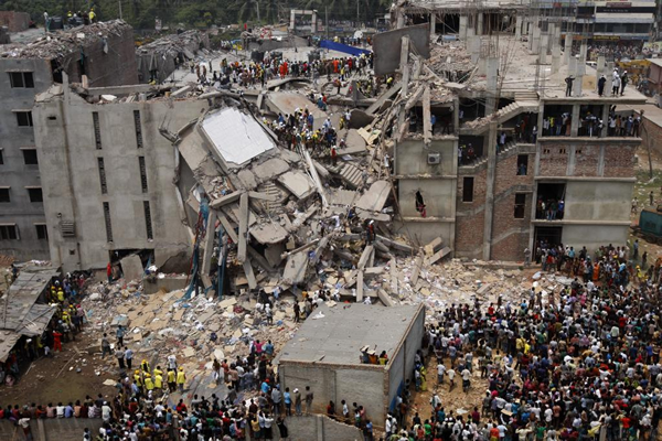 This is a photograph taken from the air or a high building of the Rana Plaza, a large 8-storey factory collapsed in the middle. There is a huge pile or rubble, dust, debris and a large crowd gathered nearby.