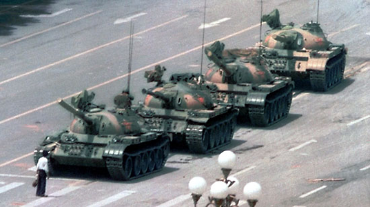 This image was taken some distance from the street. It shows a man, holding a bag, standing in the middle of the road. His feet are planted, suggesting that he has no intention of moving. About 10 feet in front of him, four tanks are slowly rolling forward. A soldier’s head can be seen just above the first tank’s hatch. He appears to be looking directly at the man on the street. Aside from the lone man and the four tanks, the street is empty.