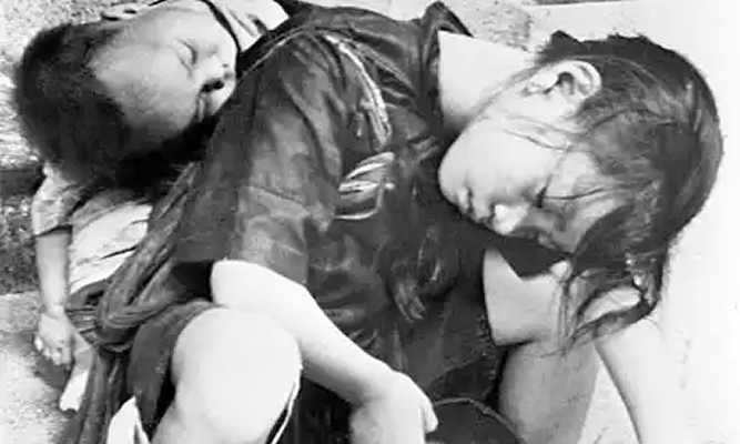 This black and white photo shows two children sleeping in awkward positions on what looks like a street corner or building stairway. Their clothes are very ragged and dirty. The young girl’s hair is disheveled. The boy, who looks like a baby, is sleeping on the girl’s back. The picture conveys a sense of extreme need.