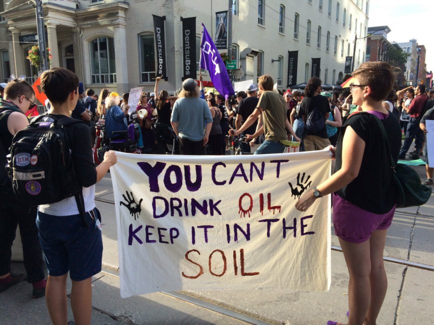 This photograph shows two protesters with their backs to the camera holding a sign that says, 'You can’t drink oil, keep it in the soil.' There are many other protesters in front of them.