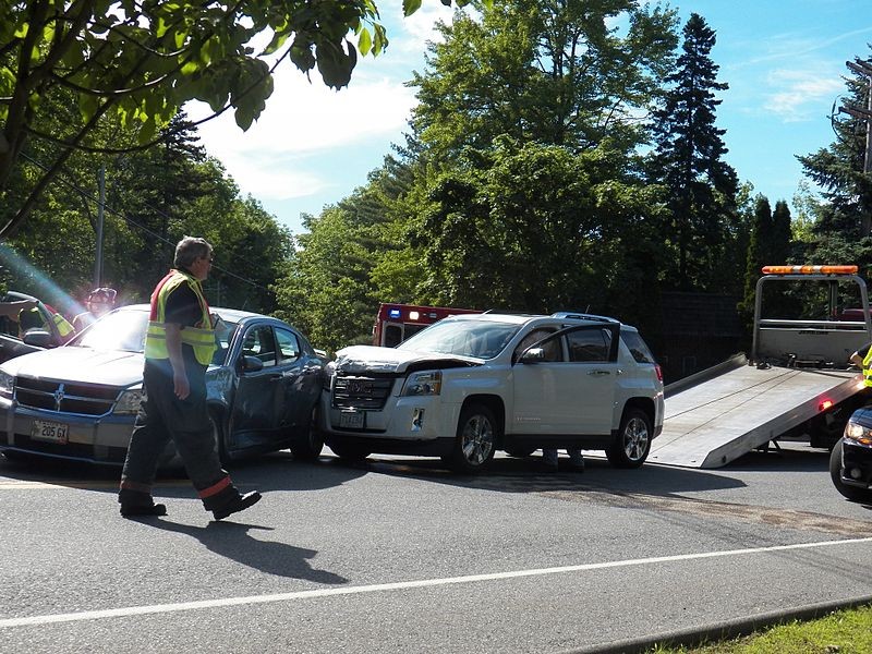 This is an image of two vehicles that have collided. There is a tow truck in the background. In the foreground a man is walking. There are emergency vehicles and personnel present in the photo.