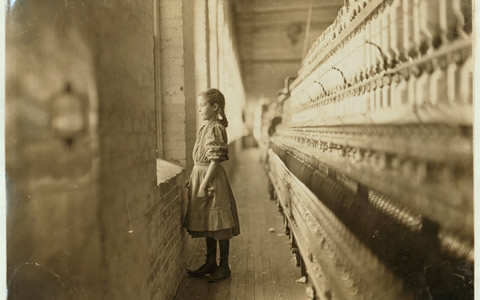 This is an image of a child standing at a window in an old factory.  The walls are made of brick. It is a black and white photo likely taken in the early years of the 20th century. She is approximately 10 years old and she is wearing an apron.
