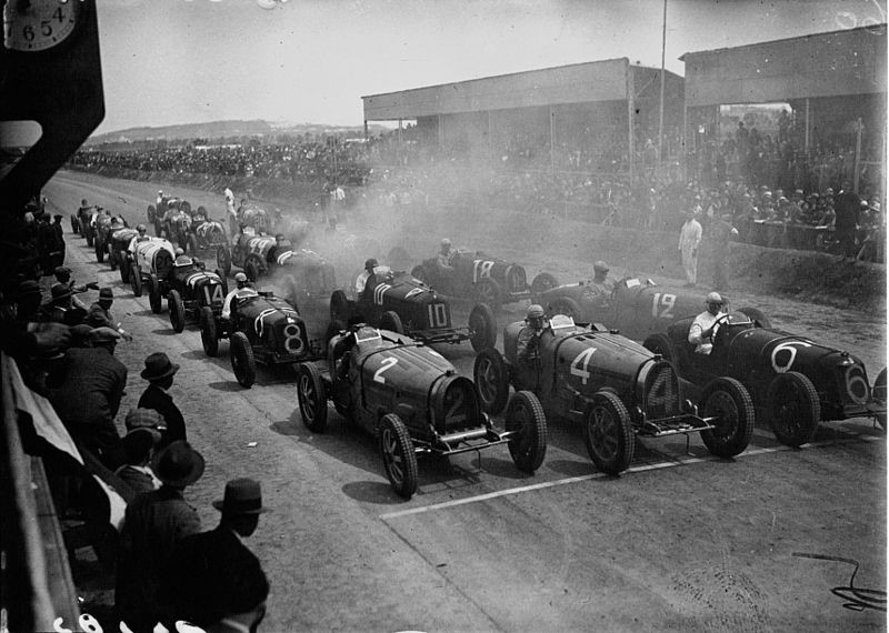 This is an image of the starting line at the 1932 Tunis Grand Prix. The photo is black and white. The cars are ready to begin the race. People are lined up on either side of the racetrack.