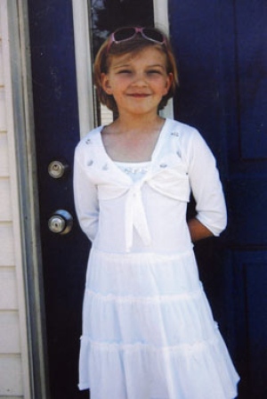 This is a photograph of a young girl in a white dress standing in front of a door. She has a big smile on her face.