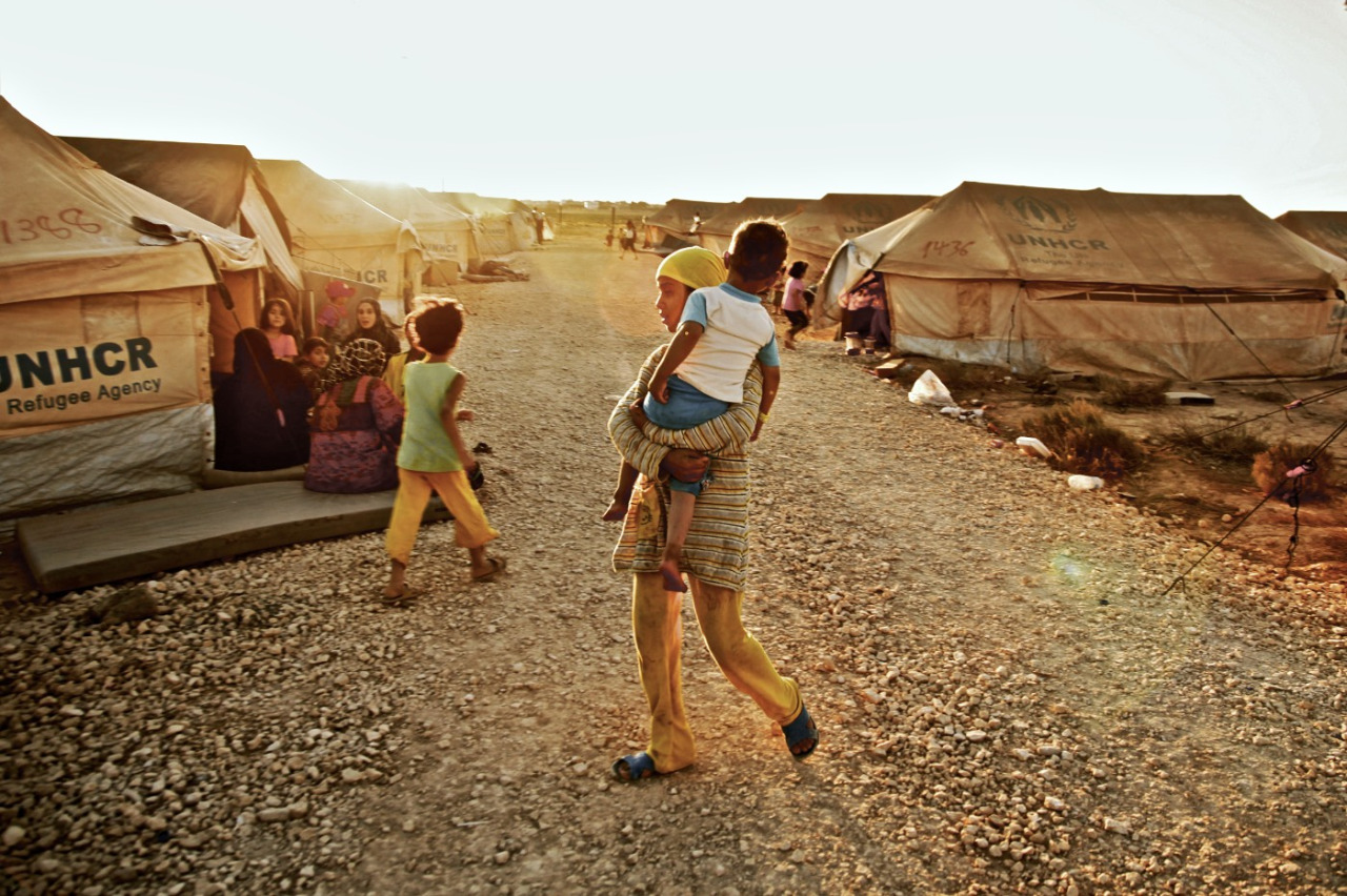 This is an image of a woman in a refugee camp. She is carrying a child. There are other people and tents in the background.
