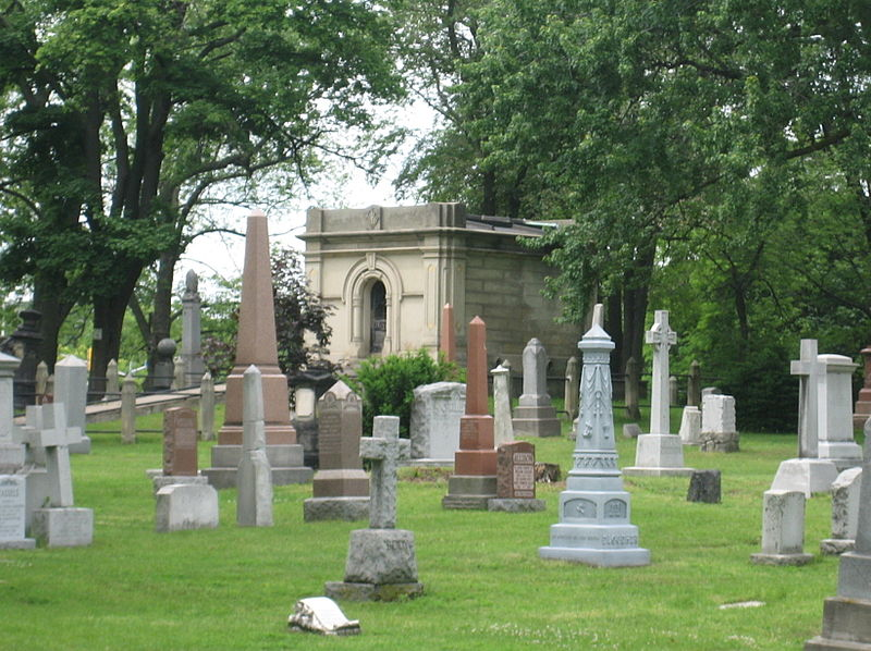 This is an image of St. James Cemetery in Toronto. There are a number of headstones of various shapes and sizes in the foreground. There is a mausoleum in the background. There are many trees in the photo also.