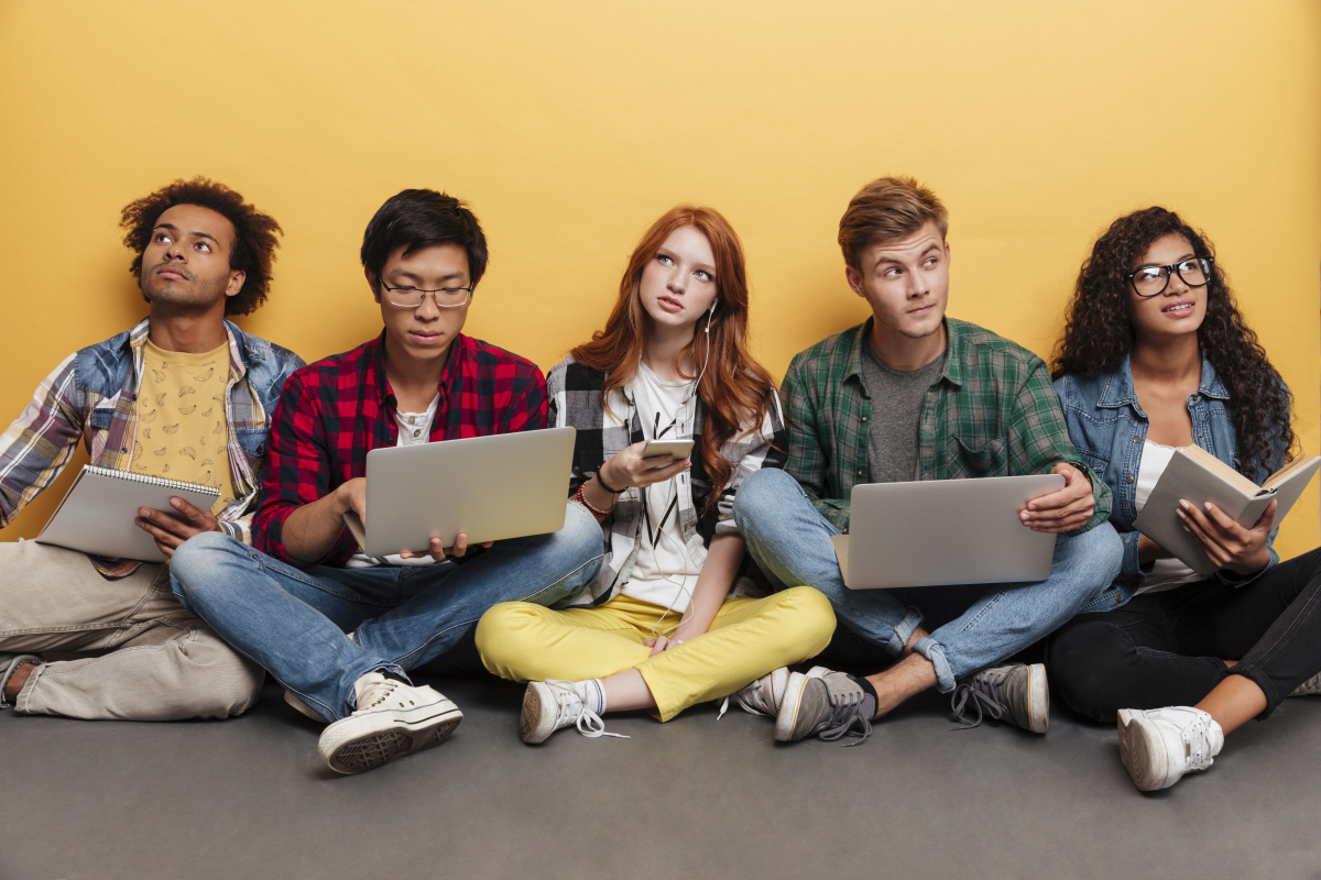 Group of young people sitting on the ground, holding a variety of laptops, notebooks, and books. 
