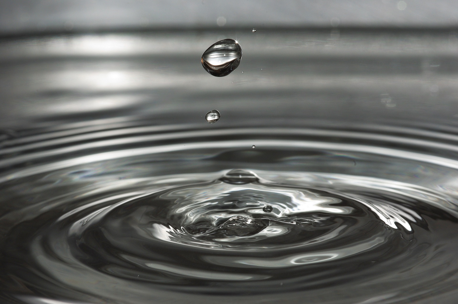 This is a black and white image of the close up of a single drop of water starting ripples in a pond.