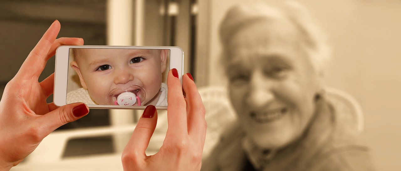 This is an image containing a close up of a smartphone that features a baby girl on the screen while in the background, there is an out of focus, sepia-coloured older woman.