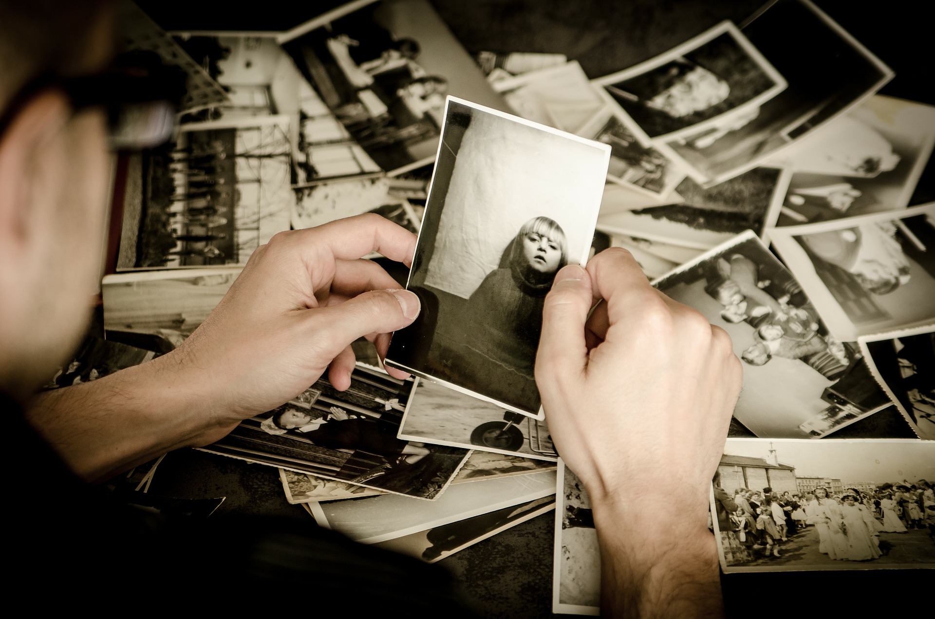 This is an image of a black and white over the shoulder shot of a hand. It is holding the picture of a young girl. A bunch of photos is scattered on the table below.