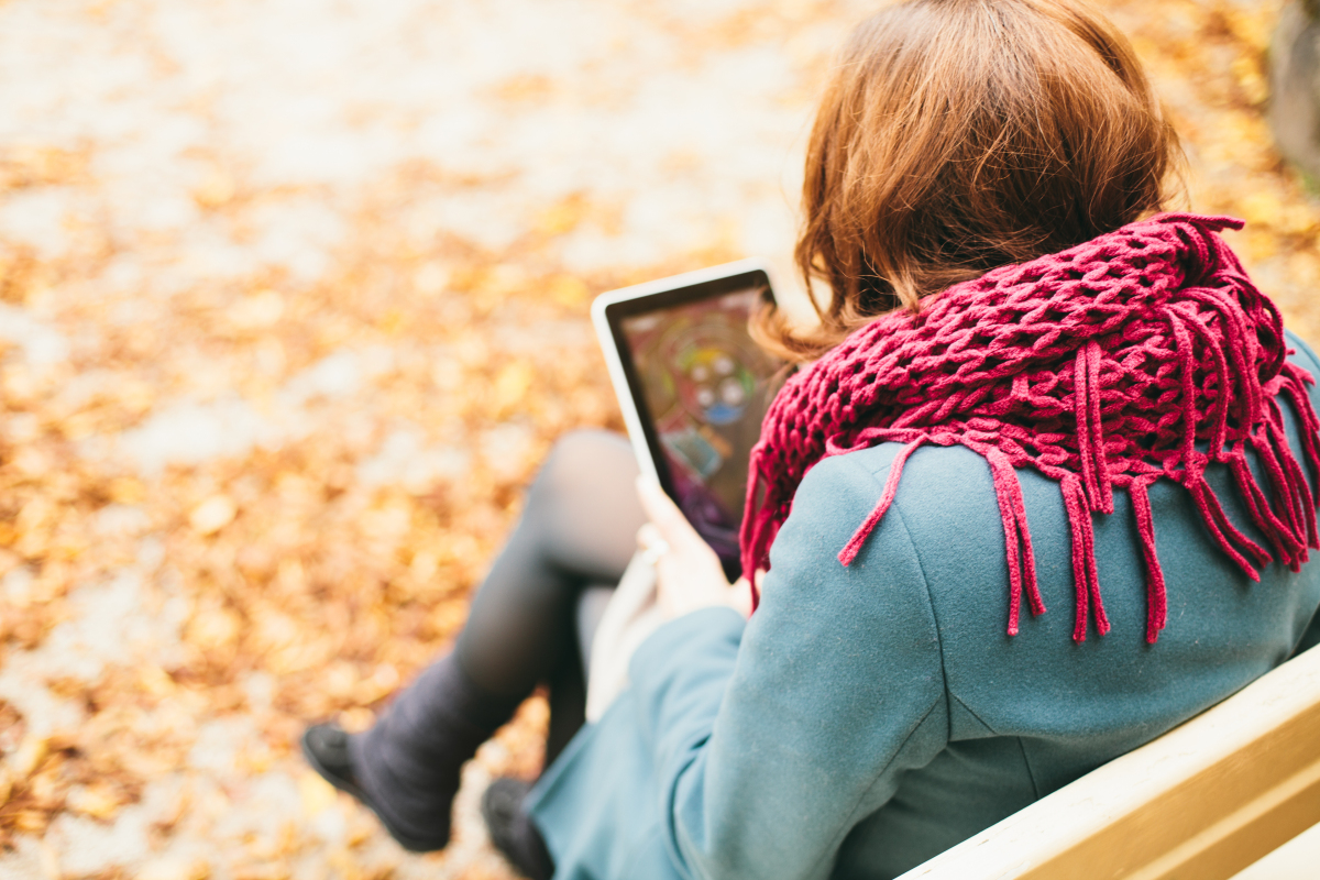 This is a photograph of a woman sitting on a bench in a park, reading something on a tablet.