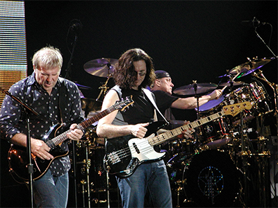 This is an image of two guitarists in the foreground with a drummer in the background.