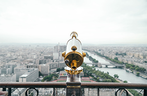 This is an image of a telescope on a railing overlooking a cityscape below.
