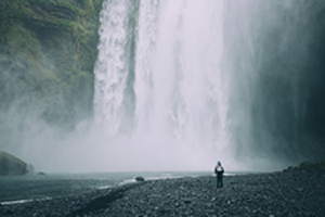 This is an image of a large waterfall pouring into a river while a hiker looks on from the shore.
