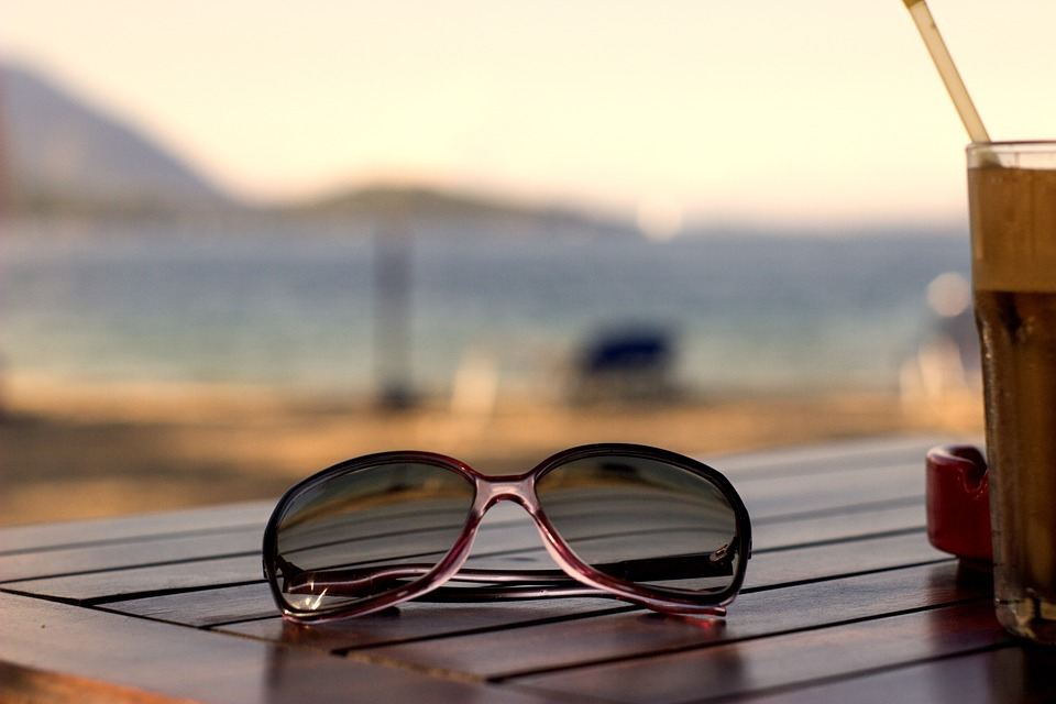 This is a close-up image of a pair of sunglasses lying on a wooden table with a blurred beach in the background.
