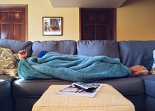 This is an image of a person lying on a couch with a soft blue blanket covering everything except their hand and feet. There is a table in front that has a remote control and magazine on it.