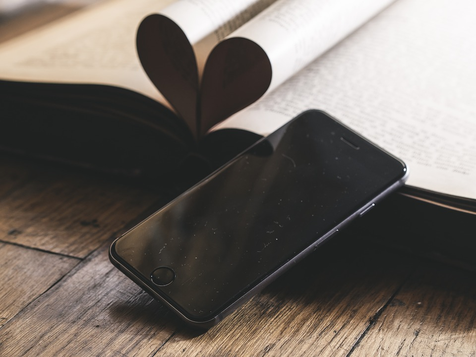 This is an image of a smart phone leaning against an open book, the pages of which are folded to make the shape of a heart.