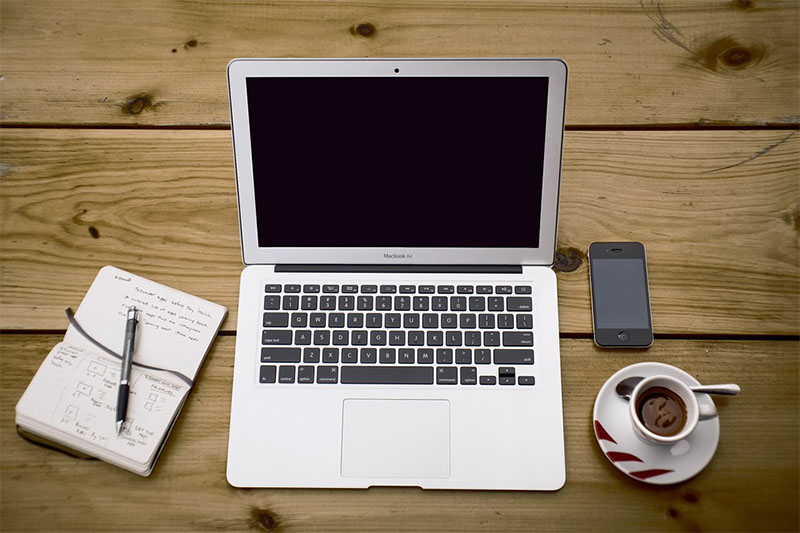 This is a photograph of a desk with a pen and notepad, laptop computer, cell phone and coffee.
