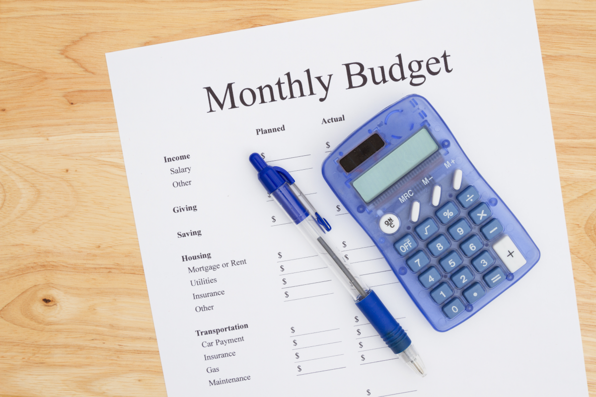 This graphic shows a print out of a monthly budget with a pen and calculator on a desk.