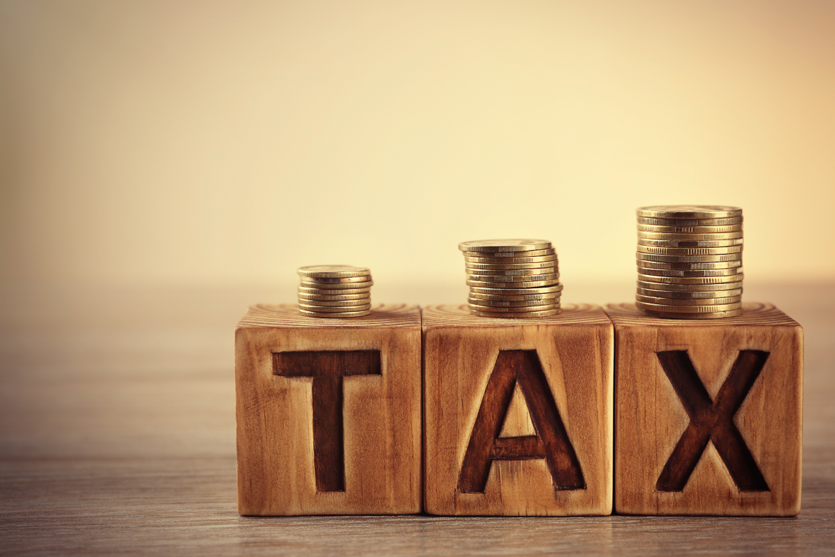 This graphic shows the word tax spelled out with wooden blocks. There are coins stacked on the wooden blocks.