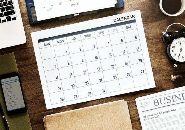 This graphic shows a blank calendar on a desk with a clock, cellphone, laptop, day planner, file folder, cup of coffee and newspaper