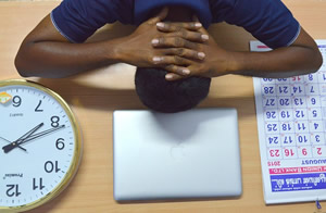 This is an image of a person with his face down on a table. His hands are clasped around the back of his head. On the table is a laptop, a calendar, and a large clock. The person appears to be stressed about his tasks and deadlines.