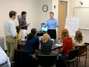 This is an image showing a group of adults in business attire sitting around a conference table while one man addresses the group