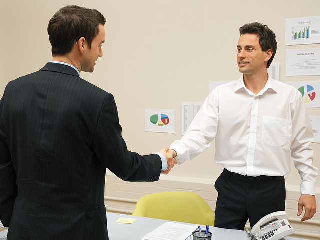 This is an image showing two adults in business attire meeting for the first time, smiling, making eye contact, and shaking one another&rsquo;s hand.